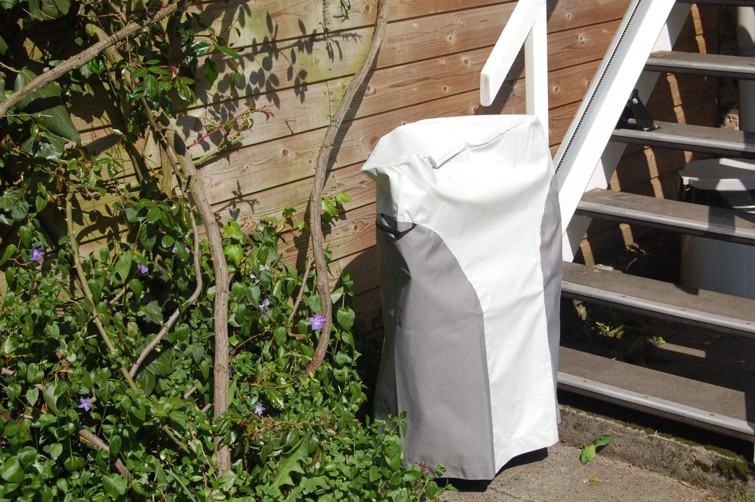 Mit Schutzhülle abgedeckter Außen-Treppenlift neben einer Holztreppe im Gartenbereich.