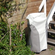 Mit Schutzhülle abgedeckter Außen-Treppenlift neben einer Holztreppe im Gartenbereich.