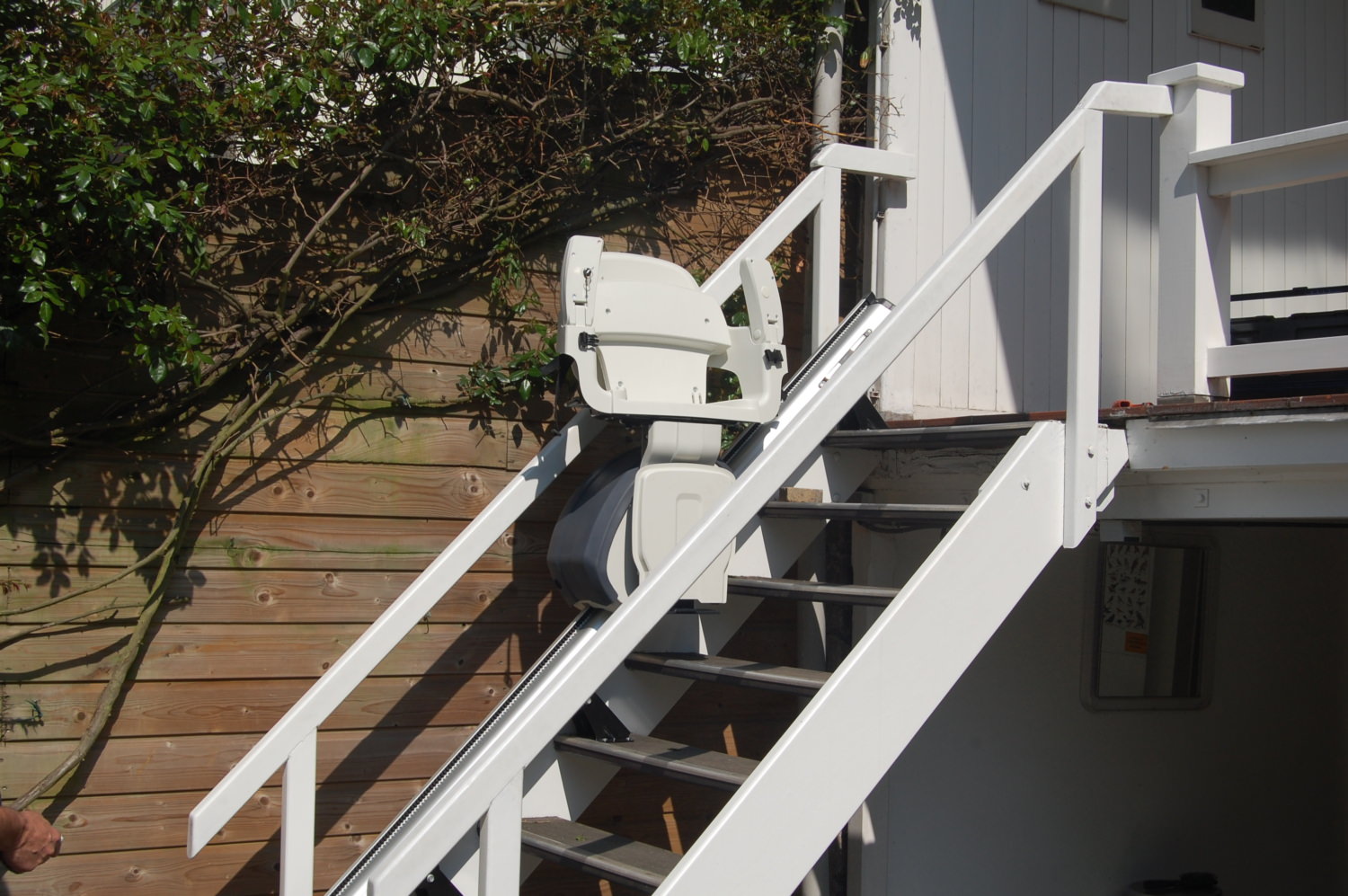Außen-Treppenlift mit weißem Sitz an einer Holztreppe im Gartenbereich installiert.