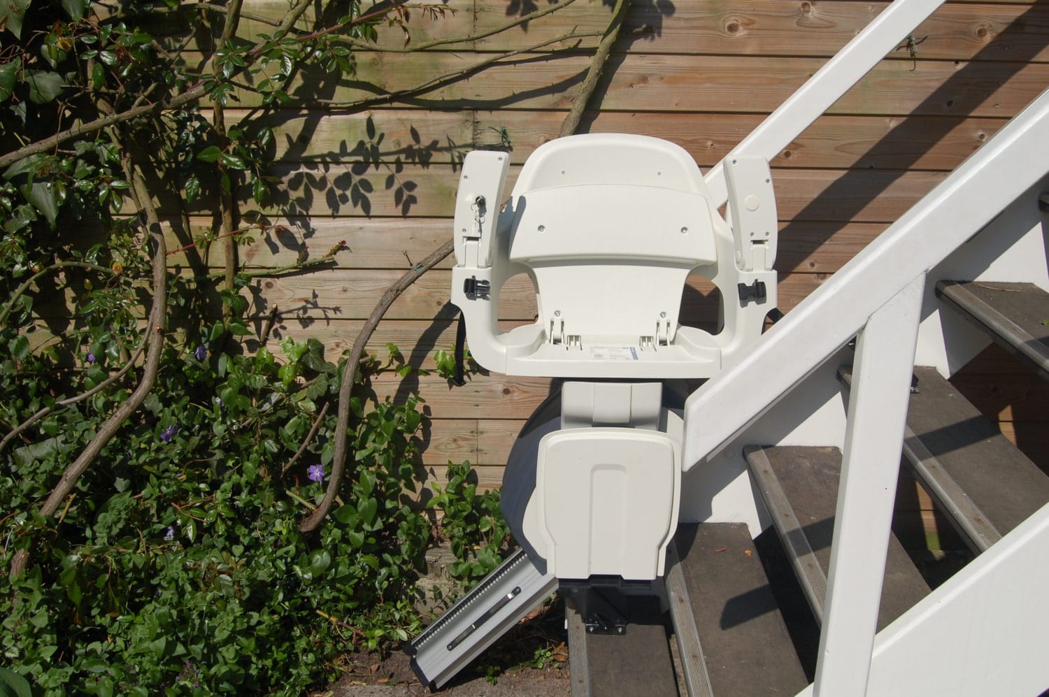 Außen-Treppenlift mit weißem Sitz an einer Holztreppe im Gartenbereich installiert.