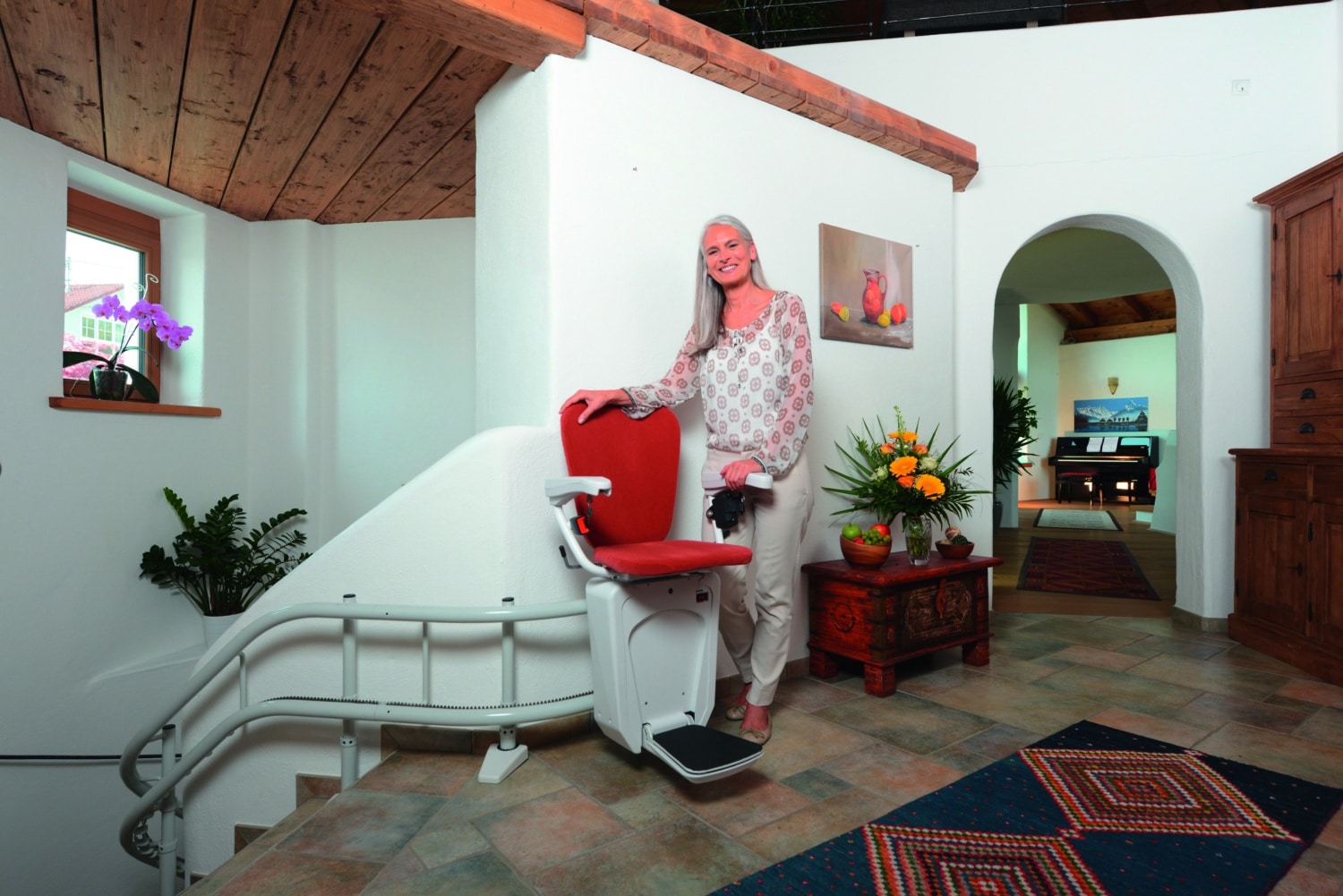 Lift Raumansicht Frau steht neben einem roten Treppenlift in einem wohnlich eingerichteten Hausflur.