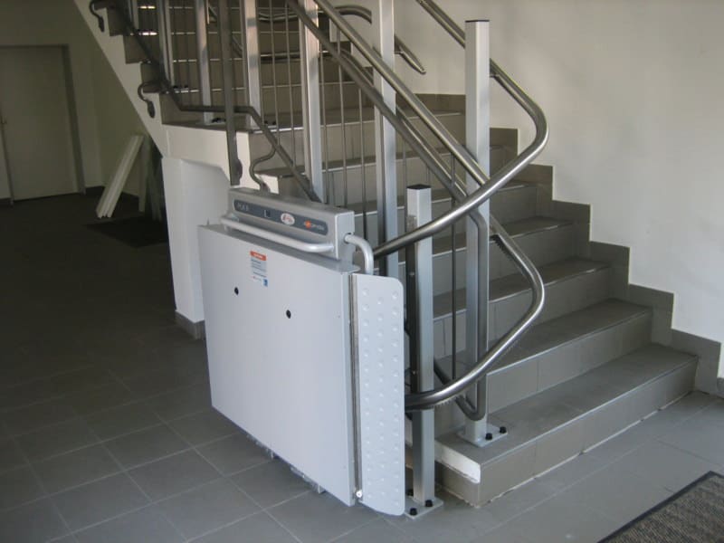 Plattformlift Plattformlift im Innenbereich entlang einer Treppe mit grauen Fliesen und Metallgeländer installiert.