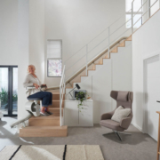 Flow-X_Main_Wide-29-scaled Seniorin fährt mit einem Treppenlift eine Holztreppe in einem modernen Wohnzimmer hinauf.