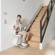 Armlehne Sitz-Treppenlift Ältere Frau sitzt auf einem Treppenlift und fährt eine moderne Holztreppe im Wohnzimmer hinauf.