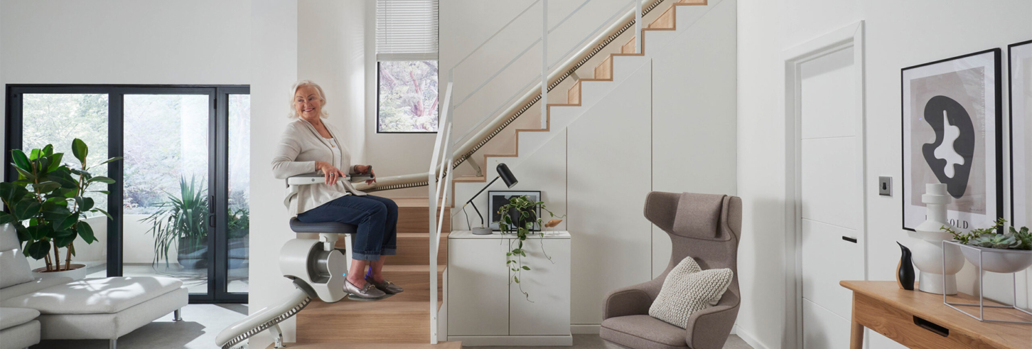 Treppenlift (kurvige Treppe) Seniorin fährt mit einem Treppenlift eine moderne Holztreppe in einem hellen Wohnzimmer hinauf.