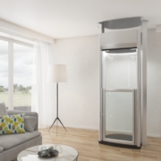 Eleganter Homelift mit verglaster Kabine in einem modernen Wohnzimmer mit Sofa, Lampe und Pflanzen.